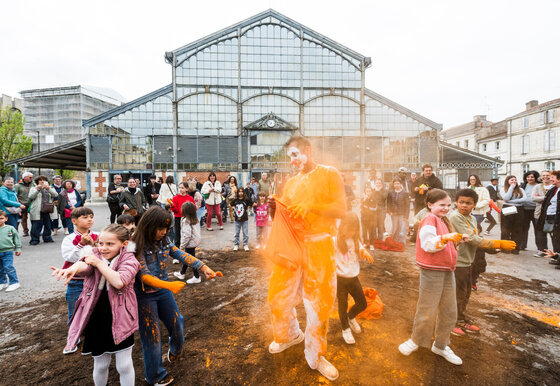 Des enfants aspergent le clown de peinture orange lors d'un spectacle de rue devant les Halles de Niort, entouré de spectateurs.