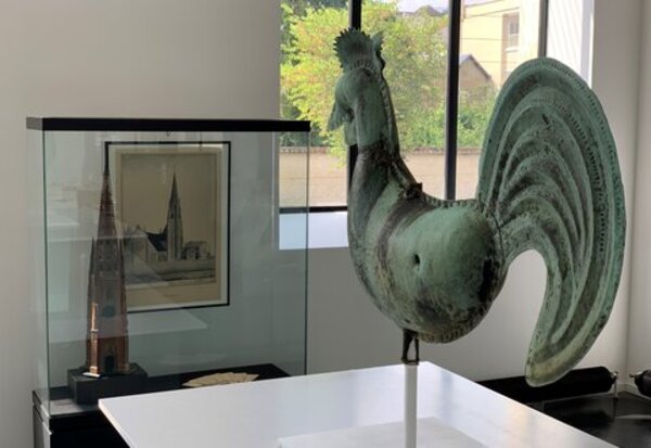Coq en cuivre à la patine verte exposé sur socle, accompagné en arrière-plan d'une vitrine présentant une maquette et un dessin de l'église