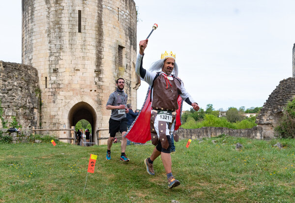 Un coureur déguisé en roi, couronne et cape rouge, brandit un sceptre devant la tour médiévale du château