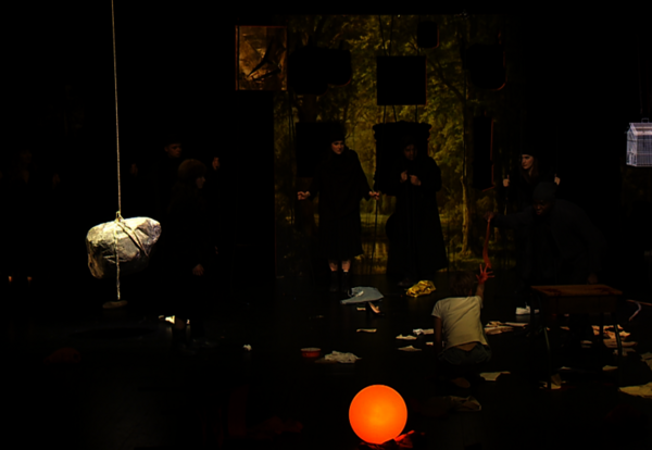 Mise en scène sombre : interprètes en robes noires sur plateau, sphère lumineuse orange au sol et décor forestier peint en fond