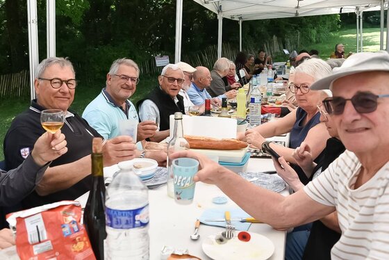 Convives souriants réunis autour d'une longue table en plein air sous un barnum, dans un parc arboré, lors des Pexinades.