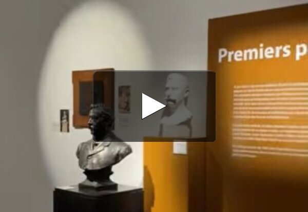 Salle d'exposition avec deux bustes sculptés, l'un en bronze et l'autre en plâtre blanc, sur socles, et un panneau orange de section
