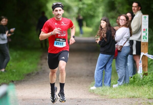 Coureur au dossard 2115 en maillot rouge progressant sur un chemin boisé, sous le regard de spectateurs alignés en bord de parcours