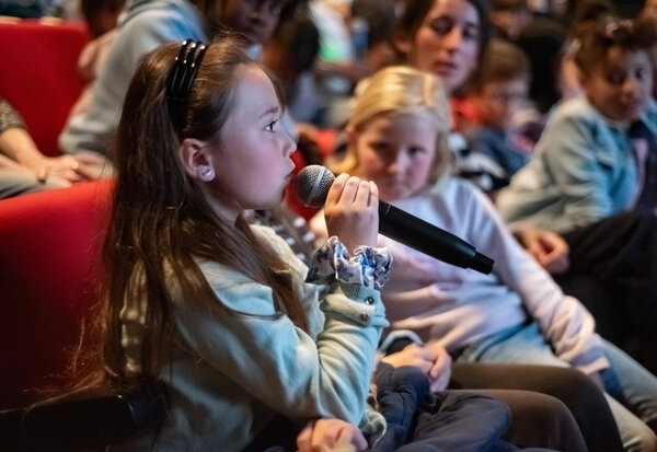 Une écolière prend la parole au microphone dans la salle de cinéma, entourée d'autres enfants et adultes