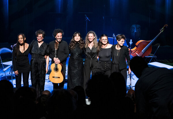 Salut final des sept artistes en tenue noire sur scène, le guitariste au centre tenant son instrument, sous des lumières bleues.