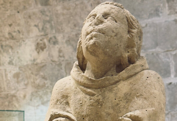 Statue médiévale en pierre d'un personnage en habit monastique, tête levée vers le ciel, dans un espace en pierre