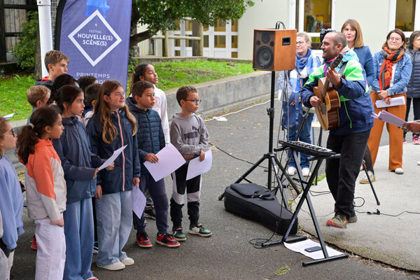 Enfants tenant des feuilles et chantant en chœur face à un musicien jouant de la guitare dans la cour de l'école
