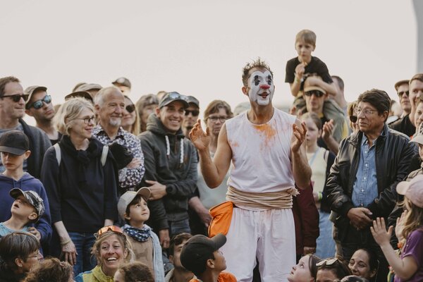 Tuga en costume blanc et maquillage de clown, jouant au cœur d'une foule de spectateurs souriants, adultes et enfants, en plein air