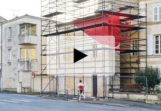 Maison Gaufrette avec échafaudage et imposante sculpture rouge en saillie sur la façade, un passant traversant au premier plan