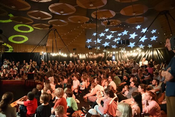 Foule de familles et enfants assis dans un chapiteau décoré de projections lumineuses en étoiles, lors d'un spectacle.