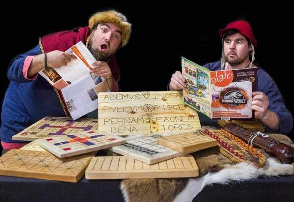 Deux comédiens en tenue médiévale, entourés de jeux anciens en bois gravé et magazines modernes, avec des expressions théâtrales étonnées