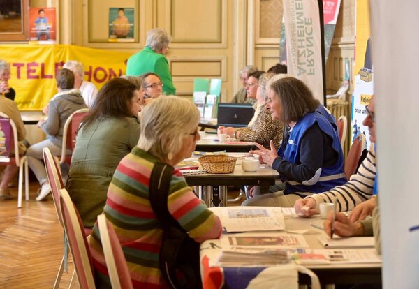 Visiteurs et représentants d'associations, dont Alzheimer France et le Téléthon, échangent autour de tables dans la grande salle.