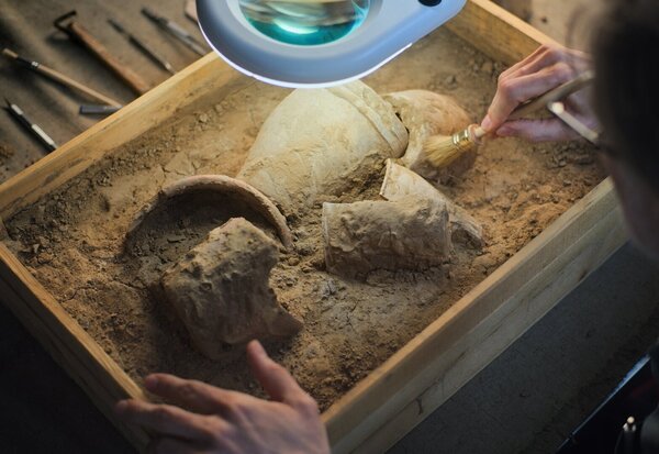 Mains brossant au pinceau des tessons de céramique dans un bac de sable, outils archéologiques disposés à l'arrière-plan