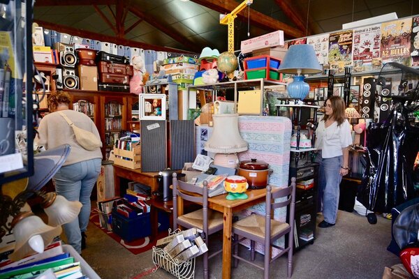 Entrepôt rempli de mobilier, luminaires, livres et objets du quotidien, avec des visiteurs qui examinent les articles disponibles.