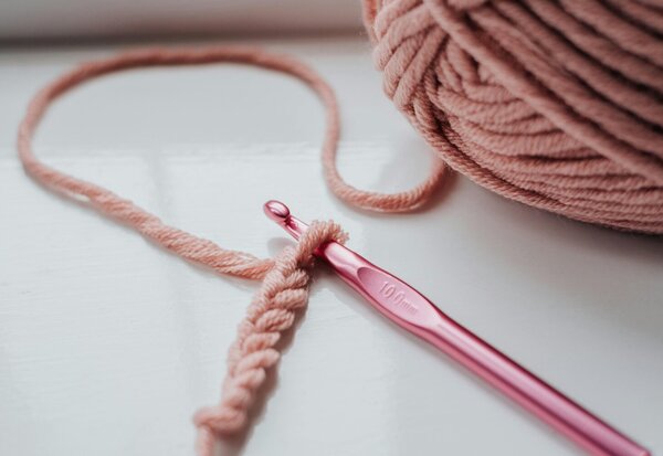 Un crochet rose métallique engagé dans une chaînette de mailles, posé près d'une pelote de fil vieux-rose