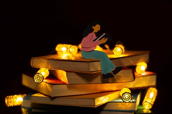 Figurine en papier lisant un livre, assise sur une pile de livres entourée de guirlandes lumineuses dorées