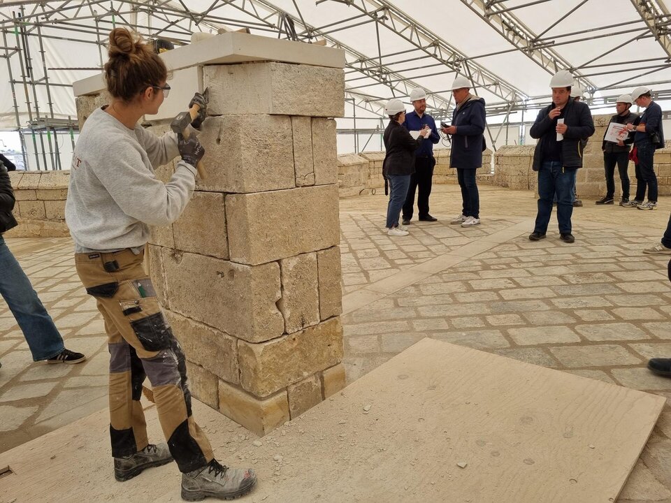 Artisane posant des blocs de calcaire sur un pilier en restauration sous tente de chantier, devant un groupe de visiteurs casqués.