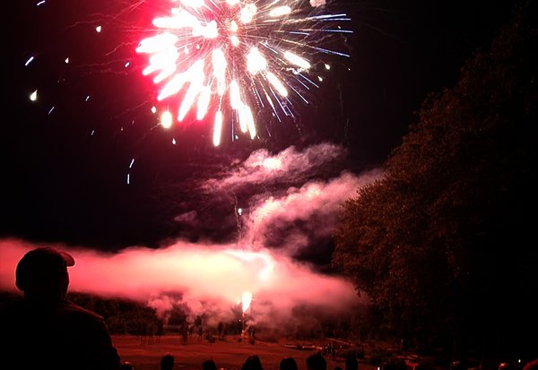 Gerbes pyrotechniques rouges et blanches éclatent dans le ciel nocturne, devant une foule de spectateurs silhouettés au premier plan.