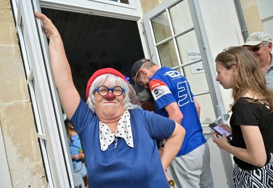 Comédienne déguisée en clown avec nez rouge et perruque blanche, accueillant des visiteurs à l'entrée d'un bâtiment
