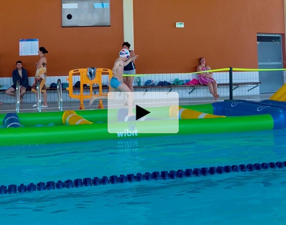 Un enfant en maillot saute et court sur les modules Wibit verts et jaunes flottant dans le bassin d'une piscine couverte.