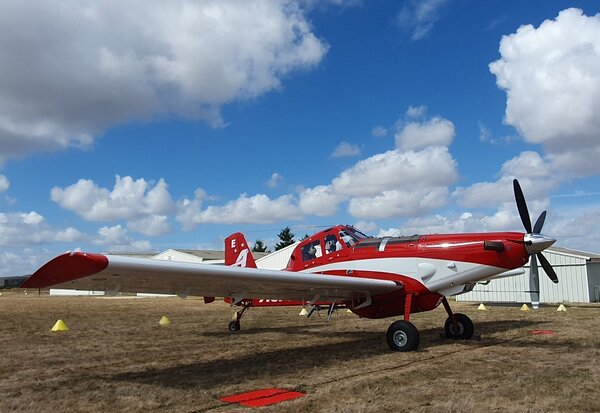 Avion biplace rouge et blanc à hélice, stationné sur une aire herbeuse, avec des hangars gris visibles en arrière-plan.