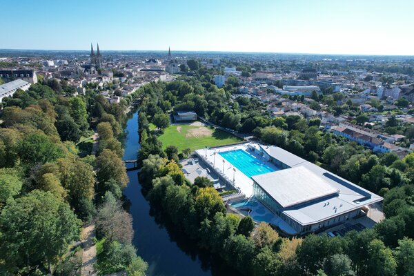 Survol aérien de la piscine Pré-Leroy, entourée de verdure et longée par la Sèvre Niortaise, avec la ville de Niort en arrière-plan.