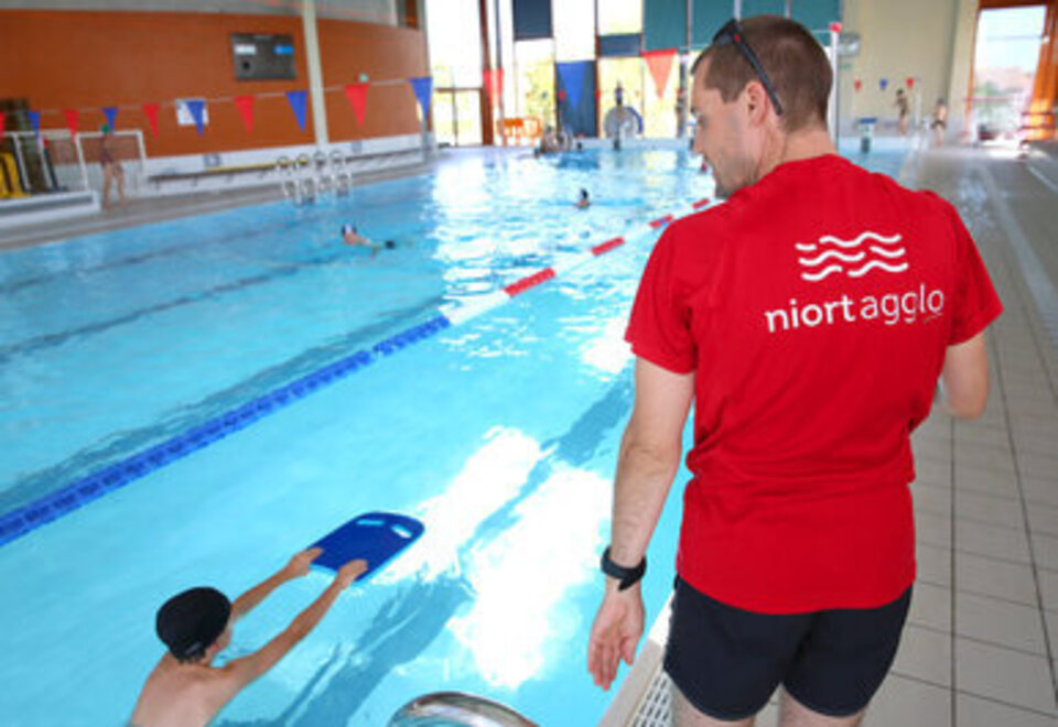 Un moniteur en t-shirt Niort Agglo surveille au bord du bassin un enfant utilisant une planche de natation.