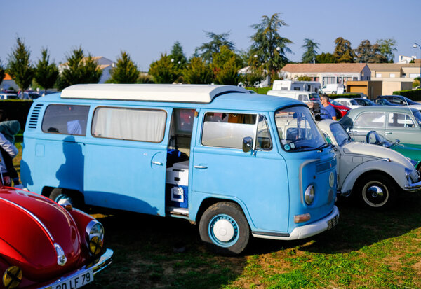 Combi Volkswagen bleu, portière coulissante ouverte, stationné entre une coccinelle rouge et une grise sur terrain herbeux