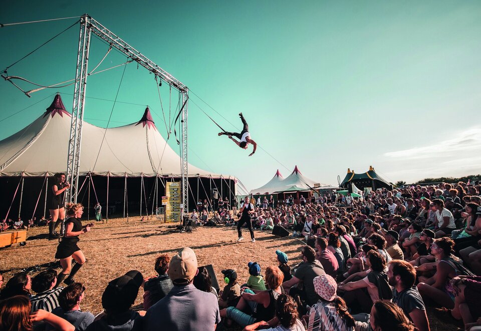 Un acrobate en plein saut périlleux aérien suspendu à une structure métallique, devant une foule nombreuse assise sous des chapiteaux de festival