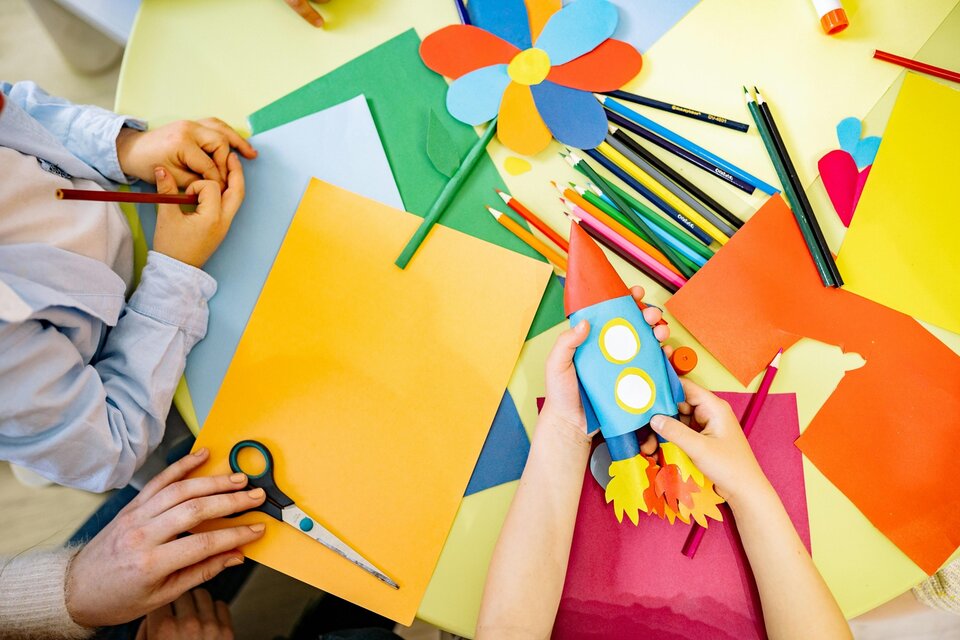 Mains créant une fusée en gobelet bleu et une fleur en papier coloré, crayons et ciseaux sur une table