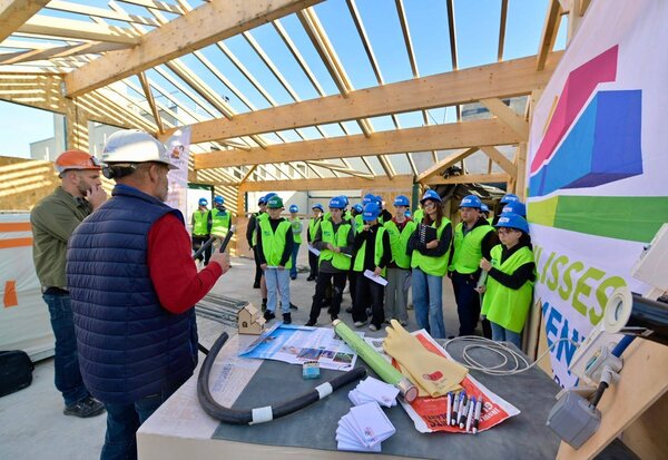 Un professionnel du bâtiment présente le chantier à un groupe d'élèves équipés de casques et gilets fluorescents.