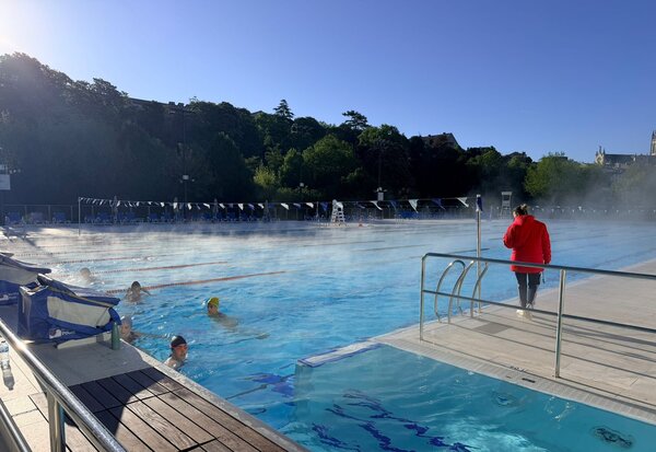 Nageuses en entraînement dans le bassin extérieur fumant sous un ciel bleu, un encadrant en veste rouge supervise depuis le bord