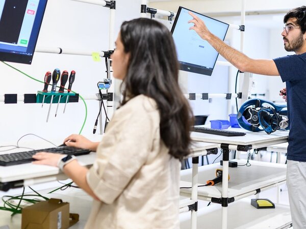Deux étudiants en ingénierie travaillent sur des postes informatiques dans un atelier équipé d'outils et d'écrans techniques.