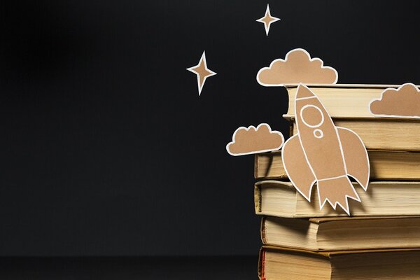 Pile de livres avec une fusée et des nuages en carton découpé évoquant l'imaginaire de la lecture enfantine