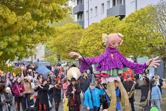 Défilé de rue animé par une grande marionnette vêtue de violet et des musiciens entourés d'une foule nombreuse