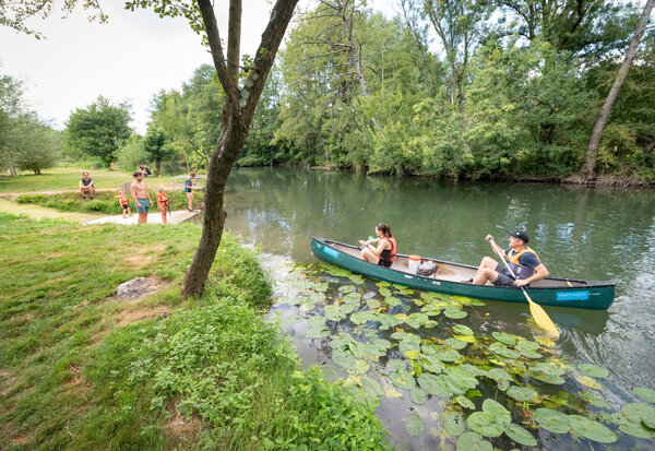 Deux personnes pagayent sur une rivière calme bordée de nénuphars, tandis que des visiteurs se regroupent sur la berge verdoyante