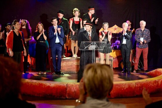Discours au pupitre dans une piste de cirque, entouré d'élus en tenue civile et d'artistes en costume de scène