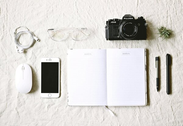 Carnet de notes ouvert, appareil photo argentique, smartphone, souris et stylos posés sur fond blanc