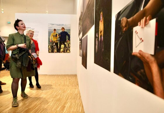 Deux visiteuses souriantes contemplent des tirages grand format accrochés sur des cimaises blanches dans une salle au parquet clair