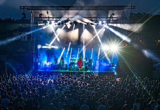 Grande scène illuminée de faisceaux bleus, artiste en tenue rouge face à une foule nombreuse lors d'un concert en plein air