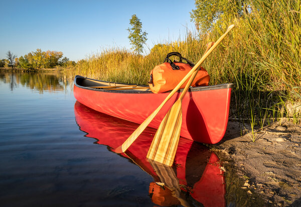 Canoë rouge avec pagaies en bois et sac étanche posé sur la rive d'une rivière calme aux reflets automnaux