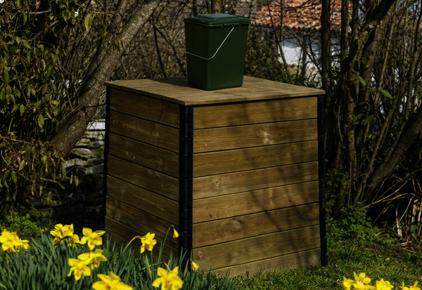 Composteur en bois avec un seau vert pour biodéchets posé dessus, installé dans un jardin fleuri de jonquilles