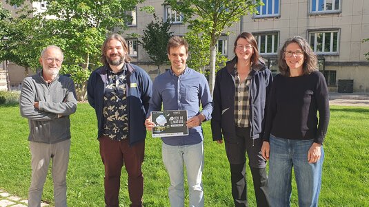 Cinq personnes posant en extérieur sur une pelouse urbaine, l'une tenant le livret du programme de l'événement