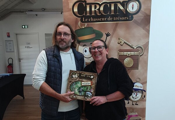 Deux personnes tenant ensemble la boîte du jeu Circino – Destination Deux-Sèvres devant le kakémono promotionnel du jeu
